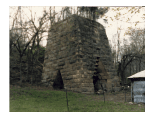 This photograph shows the abandoned Oak Ridge charcoal furnace. The furnace, located in Lawrence County, Ohio, was built in 1856 by William Williams Mather and O.M. Mitchell. Mather was the first state geologist of Ohio. Mather and Mitchell failed to make a profit and soon sold the furnace. It was later acquired by Hecla furnace. The photograph, which measures 3.5 by 4.5 inches (8.89 by 11.43 cm), was taken by Virgil and Marguerite Ramsey.  The furnace was one of many in the Hanging Rock Iron Region, an area rich in red hematite iron ore and the other materials needed to make iron, limestone and charcoal. The region includes the Ohio counties of Lawrence, Jackson, Scioto, and Vinton as well as northeastern Kentucky and the western part of West Virginia.   Charcoal furnaces were constructed of two layers, an outer one of sandstone blocks and an inner layer of fire brick or sandstone. To make a ton of iron, approximately 200 bushels of charcoal, 5,000 pounds of iron ore, and 300 pounds of limestone were heated until molten. Impurities floated to the top, forming a waste product called slag. Iron collected in the bottom of the furnace and was poured into troughs to cool. Because the troughs resembled a sow suckling her pigs, the product was known as pig iron.